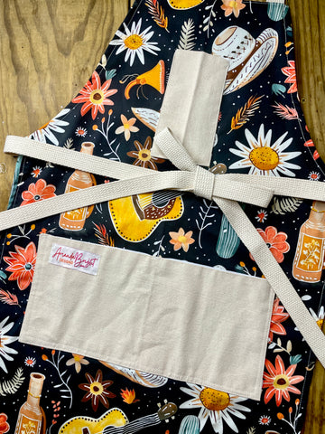 Amanda Bright Designs Desert Cowgirl Apron with colorful western guitar cactus cowboy hat sunflower pattern on black background