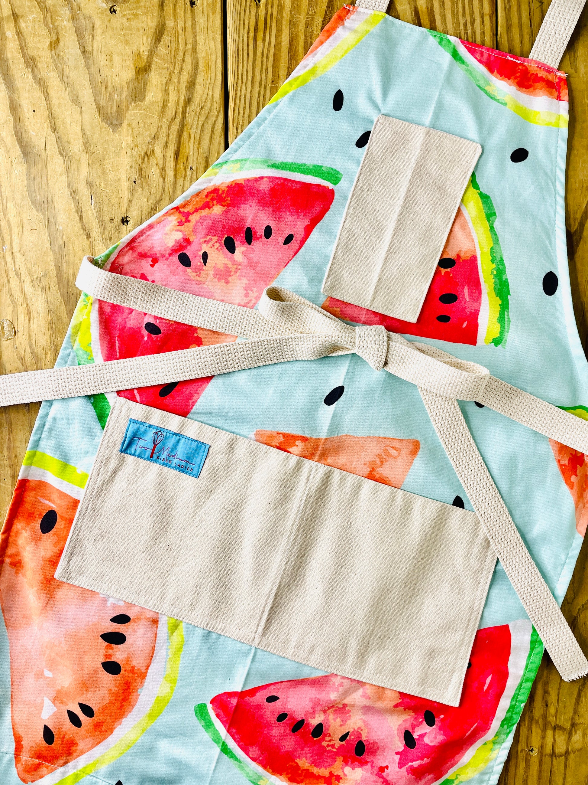 A colorful apron featuring a repeating pattern of large slices of seeded watermelon with the rind, on a pleasing mint green background. 