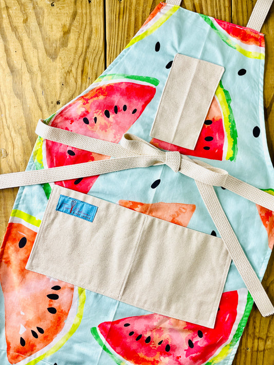 A colorful apron featuring a repeating pattern of large slices of seeded watermelon with the rind, on a pleasing mint green background. 