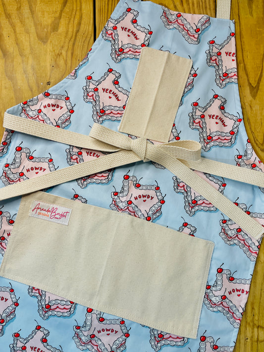Amanda Bright Designs Texas Cake Couture Apron featuring state of texas in cake form with words yeehaw and howdy in icing with cherries on baby blue background