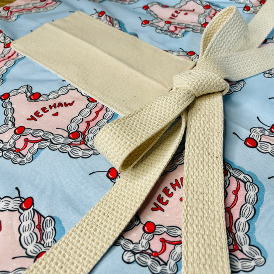 Amanda Bright Designs Texas Cake Couture Apron featuring texas shaped like a cake with pink icing cherries and words yeehaw and howdy written on the cake on a baby blue background