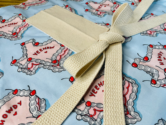 Amanda Bright Designs Texas Cake Couture Apron featuring texas shaped like a cake with pink icing cherries and words yeehaw and howdy written on the cake on a baby blue background