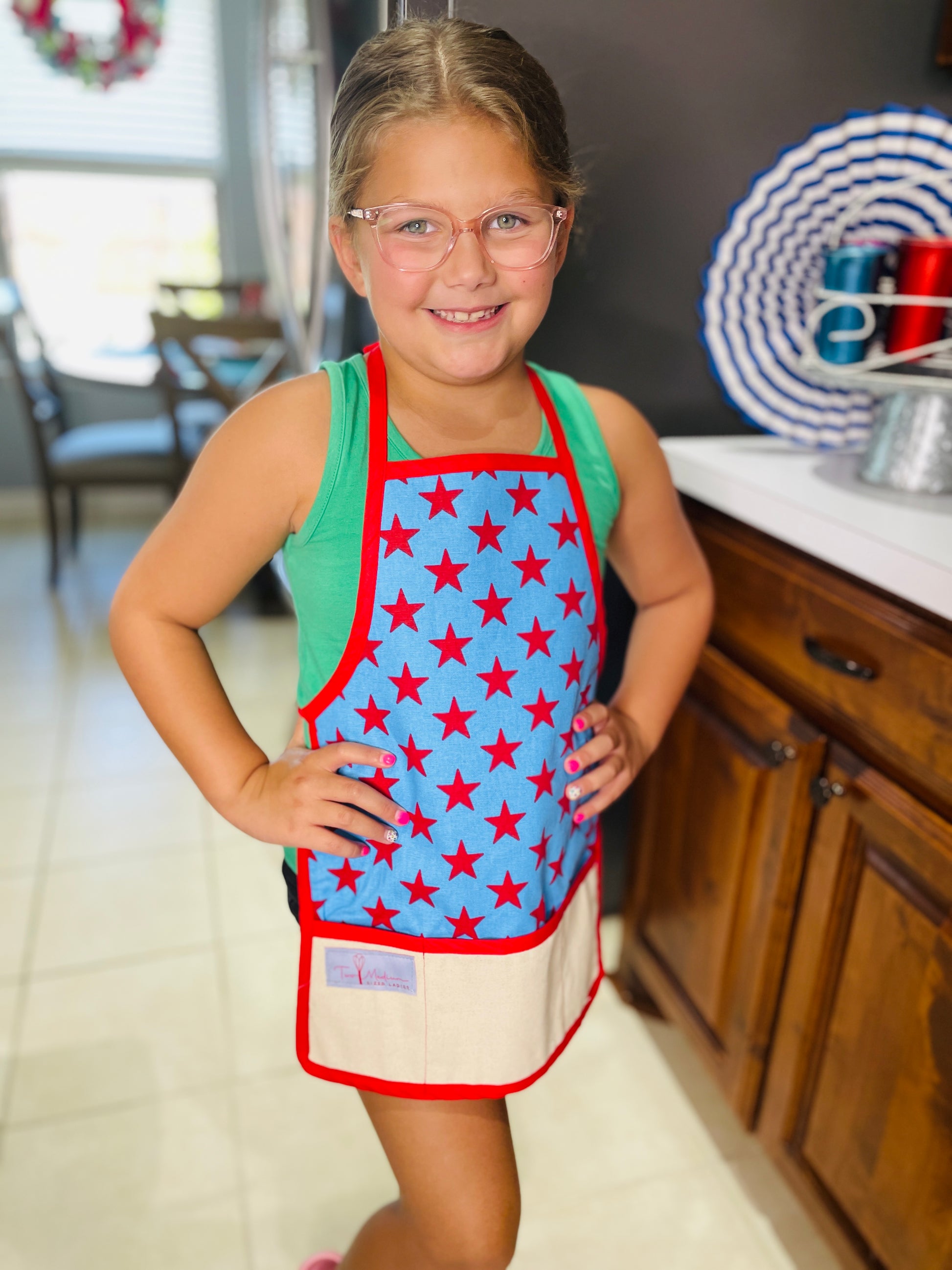 Amanda Bright Designs A child wearing a blue apron with red stars and trim, standing in a kitchen setting.
