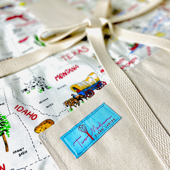 Two Medium Sized Ladies Sadie kitchen apron 50 states and their capitals historic industries road trip American heartland travel Conestoga wagon oil well longhorn airplane canoe cheese log cabin closeup view of apron