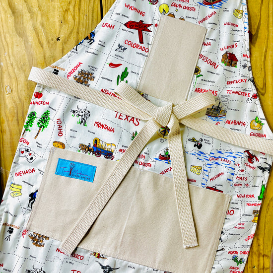 Two Medium Sized Ladies Sadie kitchen apron 50 states and their capitals historic industries road trip American heartland travel Conestoga wagon oil well longhorn airplane canoe cheese log cabin apron